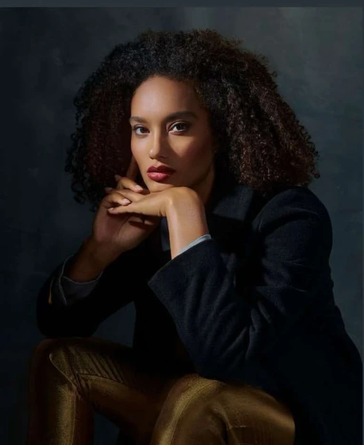 Portrait of a woman with curly hair in a black coat and gold pants, seated and posing in a studio with soft lighting.